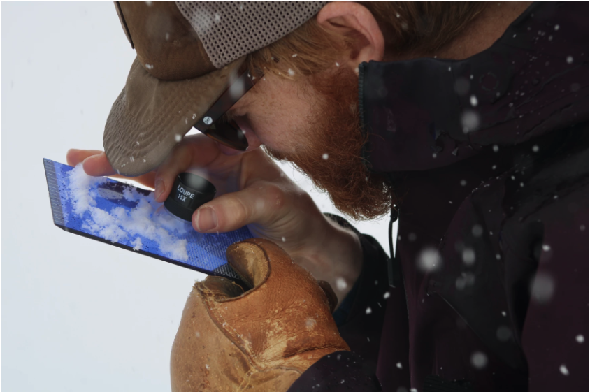 UW graduate student Danny Hogan squints into a small handheld microscope placed over a small blue board with snow on it held in his hand.