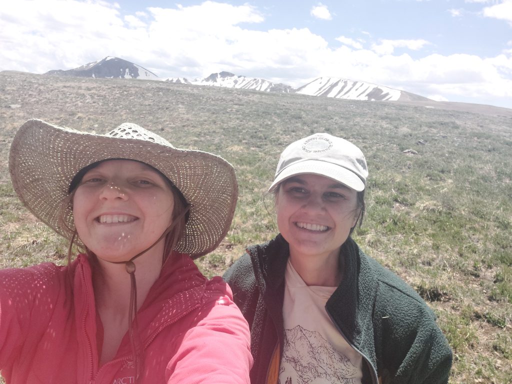 Two women in hats and outdoor clothing smiling on a mountain top