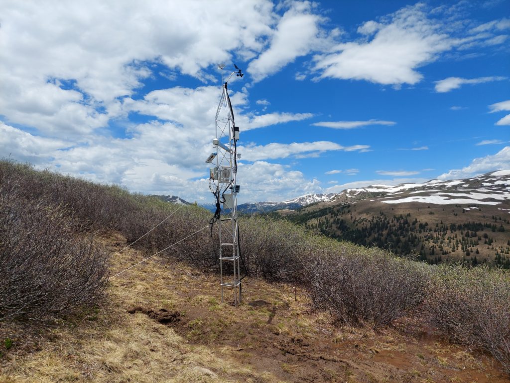 A metal tower with scientific sensors on it against the backdrop of a mountain top