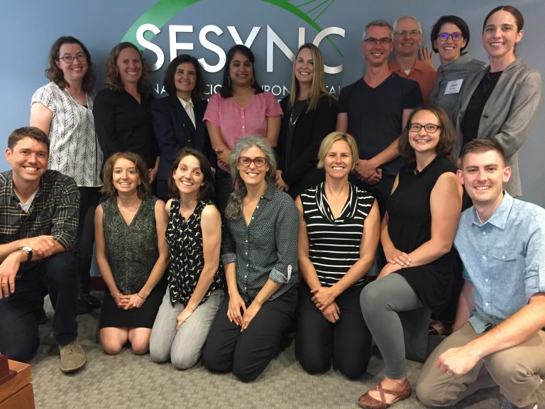 Group of researchers pose in front of National Socio-Environmental Synthesis Center (SESYNC) logo